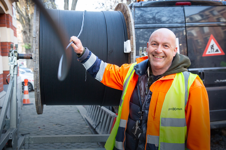 Photo d'un chantier de construction de rue. Un ouvrier chauve portant une veste fluorescente tire un câble d'un enrouleur tout en souriant à la caméra.